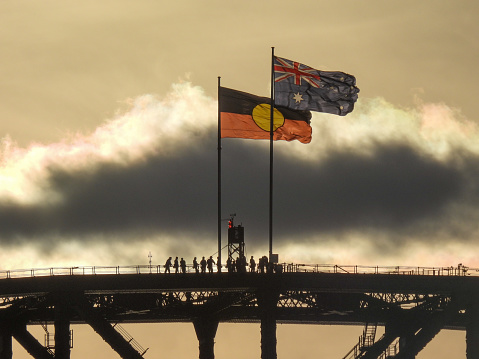 The Australian and Australian Aboriginal flags fly on the Sydney Harbour Bridge on a windy and sunny afternoon in winter.   A group of people are climbing the bridge.  This image was taken from Mrs Macquarie's Chair on 1 July 2023.