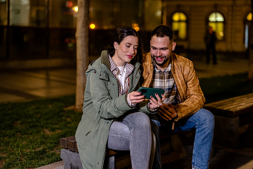 A young couple is sitting on a bench on the street at night, sharing a phone. They are deeply engrossed in whatever they are viewing and seem to be having a good time