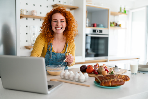 Cheerful young adult woman looking at camera while searching food recipes on laptop in the kitchen.