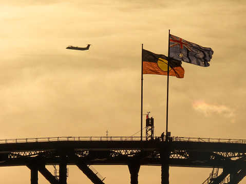 The Australian and Australian Aboriginal flags fly on the Sydney Harbour Bridge on a windy and sunny afternoon in winter.  In the background is the silhouette of a QantasLink De Havilland Canada Dash 8-400 plane, registration VH-QOX, on her landing approach to Sydney Kingsford-Smith Airport as flight QLK65D from Ballina.  A group of people visible at the base of the flags.  This image was taken before sunset from Mrs Macquarie's Chair on 1 July 2023.