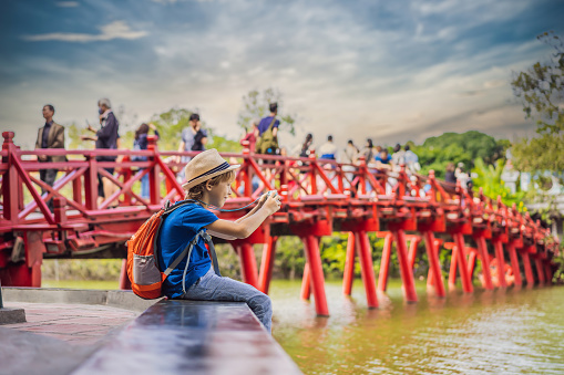 Caucasian boy tourist on background of Red Bridge in public park garden with trees and reflection in the middle of Hoan Kiem Lake in Downtown Hanoi. Traveling with children concept.