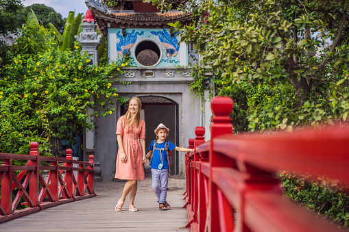 Caucasian mom and son travelers on background of Red Bridge in public park garden with trees and reflection in the middle of Hoan Kiem Lake in Downtown Hanoi. Traveling with children concept.