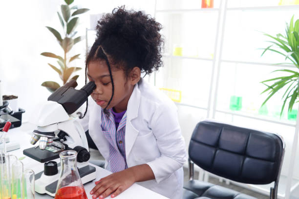 Primary school African curly hair girl looks under microscope, does science experiment in laboratory, cute scientist kid use lab equipment to learn biologics chemistry in classroom, child education. la niña africana de cabello rizado de la escuela primaria mira bajo el microscopio, hace experimentos científicos en el laboratorio, un lindo niño científico usa equipos de laboratorio para aprender química biológica en el aula, educación infantil.