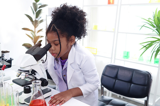 Primary school African curly hair girl looks under microscope, does science experiment in laboratory, cute scientist kid use lab equipment to learn biologics chemistry in classroom, child education.