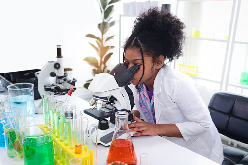 Primary school African curly hair girl looks under microscope, does science experiment in laboratory, cute scientist kid use lab equipment to learn biologics chemistry in classroom, child education.