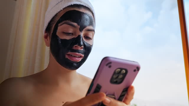 Slow motion of Hispanic young on the phone while drying up a peel off charcoal facial cleansing mask. Looking outdoors at the bedroom window