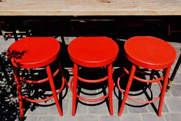 bright red wood bar stools in closeup view under bar table. exterior terrace space. gray concrete tile floor. strong shadows and contrast. bright lights. restaurant and hospitality. outdoor terrace scene.