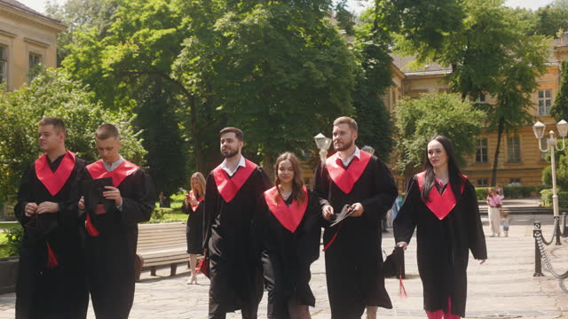 Graduating students dressed in gowns and graduation caps walk together, talking and enjoying themselves. There is a cheerful atmosphere among them on a sunny summer day.