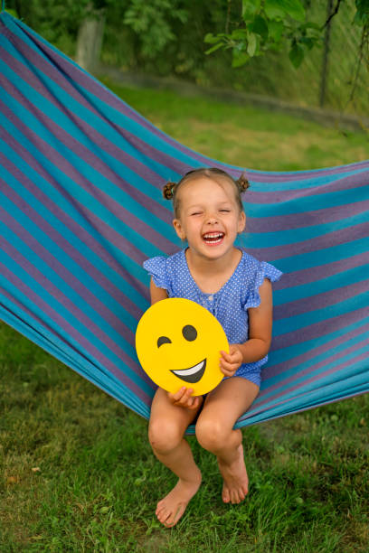 World emoji Day used for social media communication Funny laughing girl is sitting on a hammock in the garden holding a winking smile face in her hands. World emoji Day used for social media communication child laughing hysterically stock pictures, royalty-free photos & images