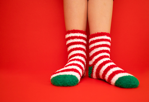 Female feet in fluffy New Year or Christmas warm socks. The colors of the socks are red and white stripes and green heels and tips. Girls legs on a red bright background. Celebration holiday concept.