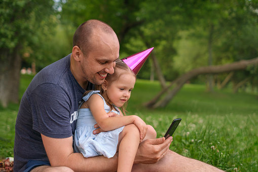 A father with little birthday girl daughter with festive hat on head communicates via video communication using smartphone. The girl accepts birthday greetings from family
