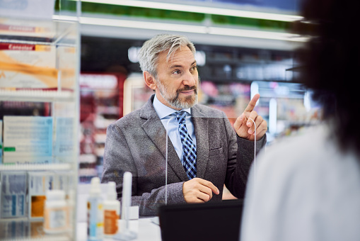 A bearded man in a suit buying medicine in a pharmacy, standing in front of the glass on a cash register.