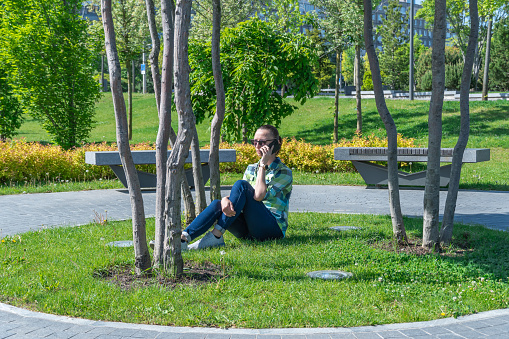 Woman sitting on park lawn and talking on mobile phone. Young adult use smart phone for chatting. Female resting and conversation on the cell phone. Leisure, technology and modern lifestyle concept.
