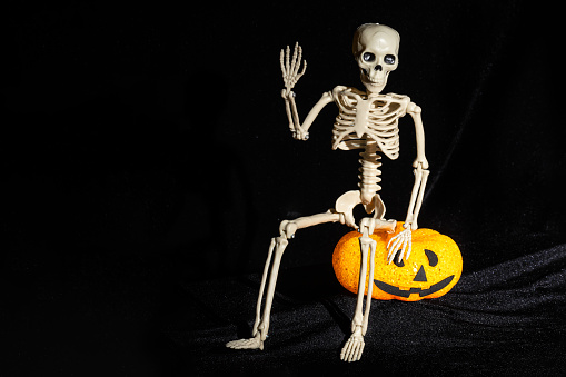Skeleton sitting on a lamp in the shape of a pumpkin with a grin and waving his hand in greeting on a black background