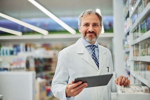 A grey-bearded male pharmacist using a digital tablet for work in the pharmacy, smiling for the camera.