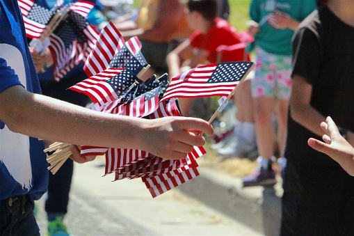 Part of human passing out American flags along parade route, hand reaching from corner of frame, blurred crowd in background. Independence Day celebration, 4th of July in small town USA.