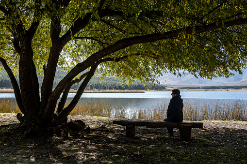 Senior woman rests sitting under a tree in front of Laguna Zeta, in Esquel, Chubut, Argentina.