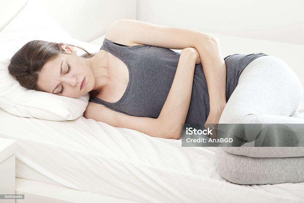 Woman lying down in bed clutching stomach with a grimace Young woman in pain lying on bed Abdomen Stock Photo