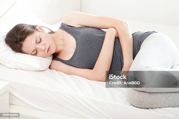 Woman Lying Down In Bed Clutching Stomach With A Grimace Stock Photo - Download Image Now