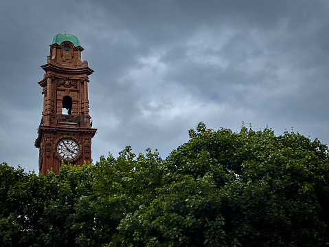 A photograph of the Victorian Kimpton Clock Tower in Manchester, England. The terracotta Grade II–listed Kimpton Clock Tower has a history that dates back to 1890. The photograph shows the Clock Tower rising from behind a row of trees and isolated against a brooding sky.