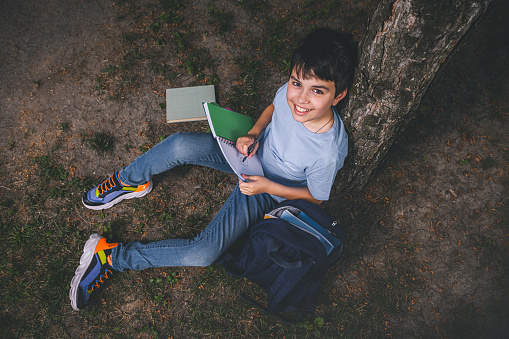 View from above of a charming adorable Caucasian smart teenage schoolboy in casual denim, doing homework, writing on copybook, sitting under tree in the park after school, smiling looking at camera