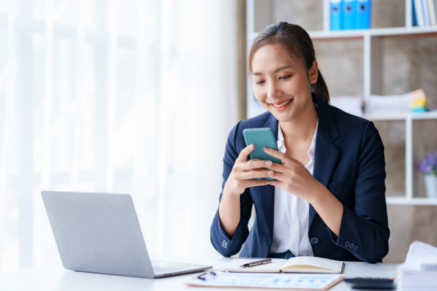Beautiful Asian businesswoman is using smartphone to chat with customers at work and play various social media applications in a relaxed smile and good mood. stock photo