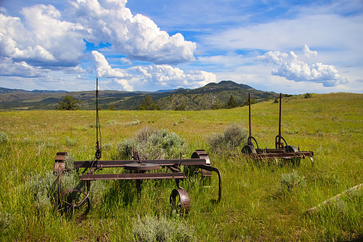 Landscape of large  clouds forming above a distant mountain on a sunny Summer day as viewed from beyond old farming equipment in a field and rolling green hills near Townsend, Montana, USA.