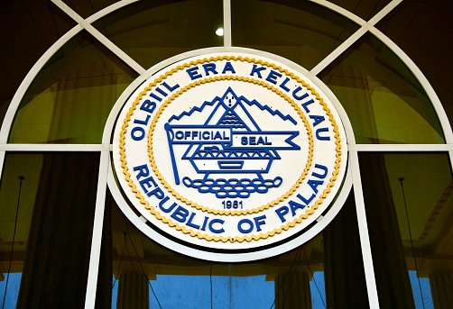 Ngerulmud, Melekeok State, Babeldaob Island, Palau: Seal of the Republic of Palau, façade of the Capitol of Palau central wing - building of the Palau National Congress, the Legislative Building (parliament). The emblem is a disk showing the traditional bai community meeting-house, standing on sixteen stones and a flag on the flagstaff reading 'Official Seal'. The year 1981 refers to the establishment of the Republic of Palau and 'Olbiil Era Kelulau' (OEK) is Palauan for “House of Whispered Decisions\
