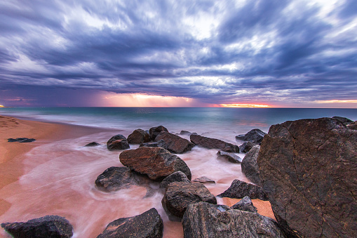 Long exposure seascape of bay of water with ominous storm clouds and lightning with smooth ocean and rocky beach coastline in Western Australia.