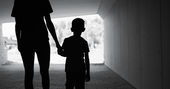 Silhouette of mother holding son hand walking city street.