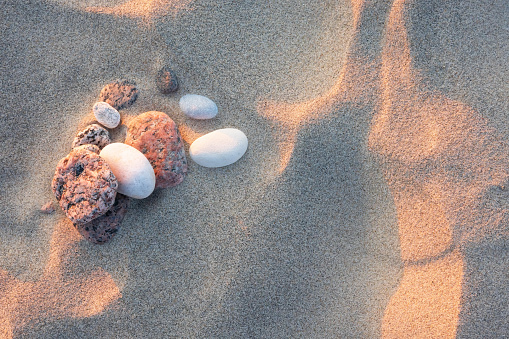 The uneven sand on the beach is illuminated by the soft light of the setting sun. There are several small sea pebbles of red and white color. Background. Texture.