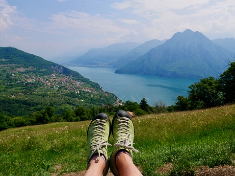 Woman's legs with green hiking boots, panoramic view of Lago d'Iseo seen from Fonteno - Riva di Solto Big Bench, Lombardy, Italy.