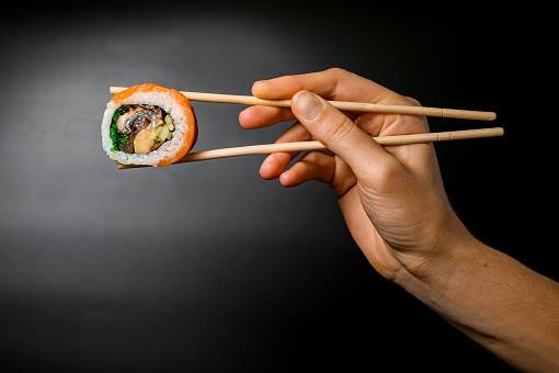 side view of one appetizing Philadelphia roll with salmon, eel, avocado, cucumber, green 
algae and cream cheese in hand holding with bamboo chopsticks on dark background