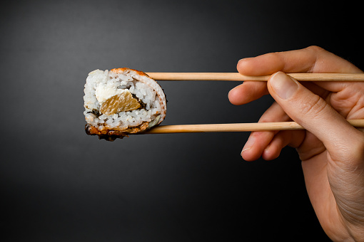 Woman's hand holds Philadelphia sushi roll with smoked eel, salmon and cream cheese with bamboo chopsticks on dark background