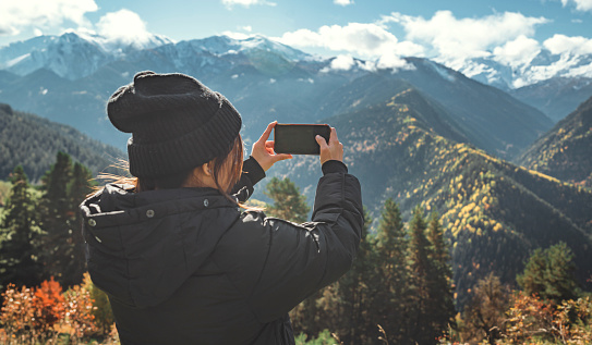 Woman traveler making photo of the snow-capped mountains with mobile smartphone, Female hands holding phone and doing nature photography while enjoying beautiful autumn nature landscape wanderlust