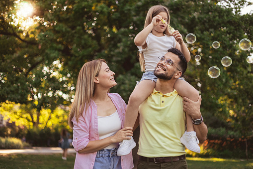 Happy Mother and Father Carrying Daughter on Shoulders as They Walk in Park Enjoying the Beautiful Day in Nature. Family and Love Concept