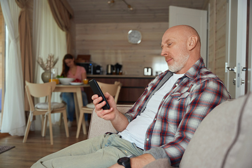 Focused male seated on sofa reading news on smartphone screen while his wife preparing food