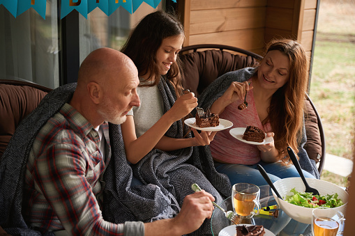 Family of three seated at festive table on cottage veranda eating chocolate cake