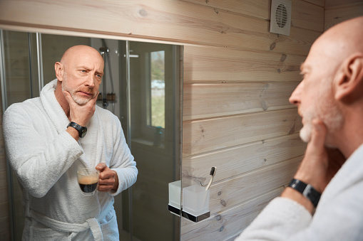 Despondent male with coffee cup in hand touching his beard in front of mirror