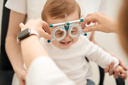 Female oculist hands putting on ophthalmic trial frame on joyous infant in parent presence