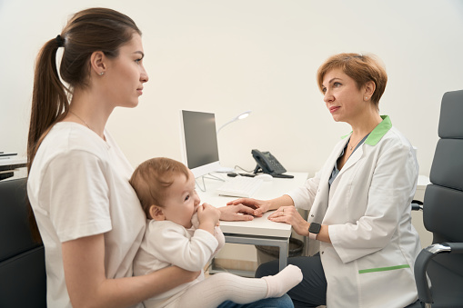 Friendly eye doctor reassuring young woman in presence of her infant during consultation