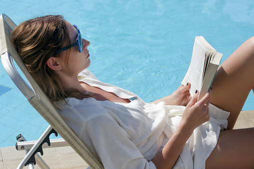 Portrait of beautiful young woman enjoying hot day reading a book next to the swimming pool. Relaxing summer vacation concept.