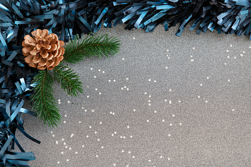 Festive frame with blue tinsel, a pine cone and fresh pine brunches, and sparkling confetti stars spread against a silver gray background, Christmas or winter holidays themed decoration with copy space.