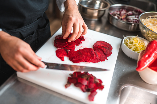 Asian employee working during daytime. Male hands are carefully cutting a beet on a cutting board, using a sharp kitchen knife.