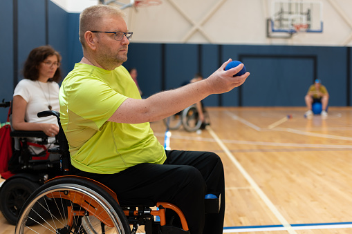 Disabled person with severe dystrophy in a wheelchair plays bocce in the gym on the floor. Wheelchair and power wheelchair.
