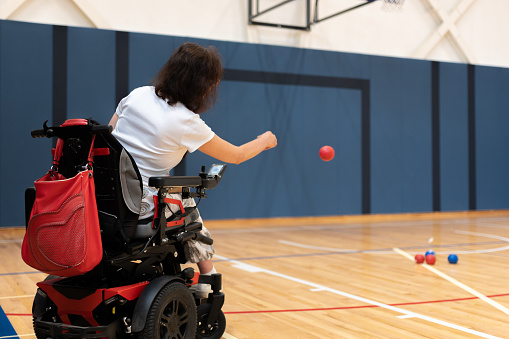 Disabled person with severe dystrophy in a wheelchair plays bocce in the gym on the floor. Wheelchair and power wheelchair.
