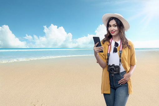 Asian woman with a hat and camera holding a mobile phone traveling on the beach with blue sky background