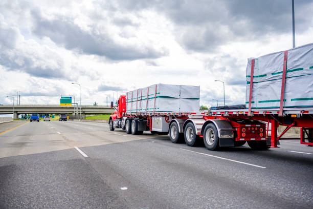 istock Bright red day cab big rig classic semi truck tractor transporting covered fastened cargo on two flat bed semi trailer driving on the wide multiline highway road with gray cloudy sky 1510208409