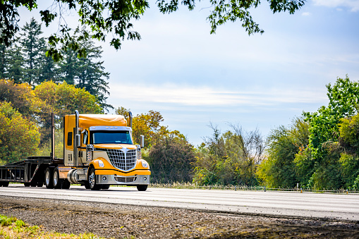 Industrial grade yellow bonnet big rig long hauler semi truck tractor with extended cab for truck driver rest transporting empty step down semi trailer driving on the highway road at sunny day