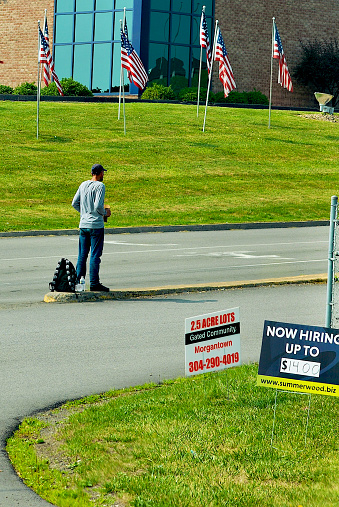 Morgantown, West Virginia, USA - June 29, 2023: A man holds a sign asking for help at an intersection in front of U.S. flags.